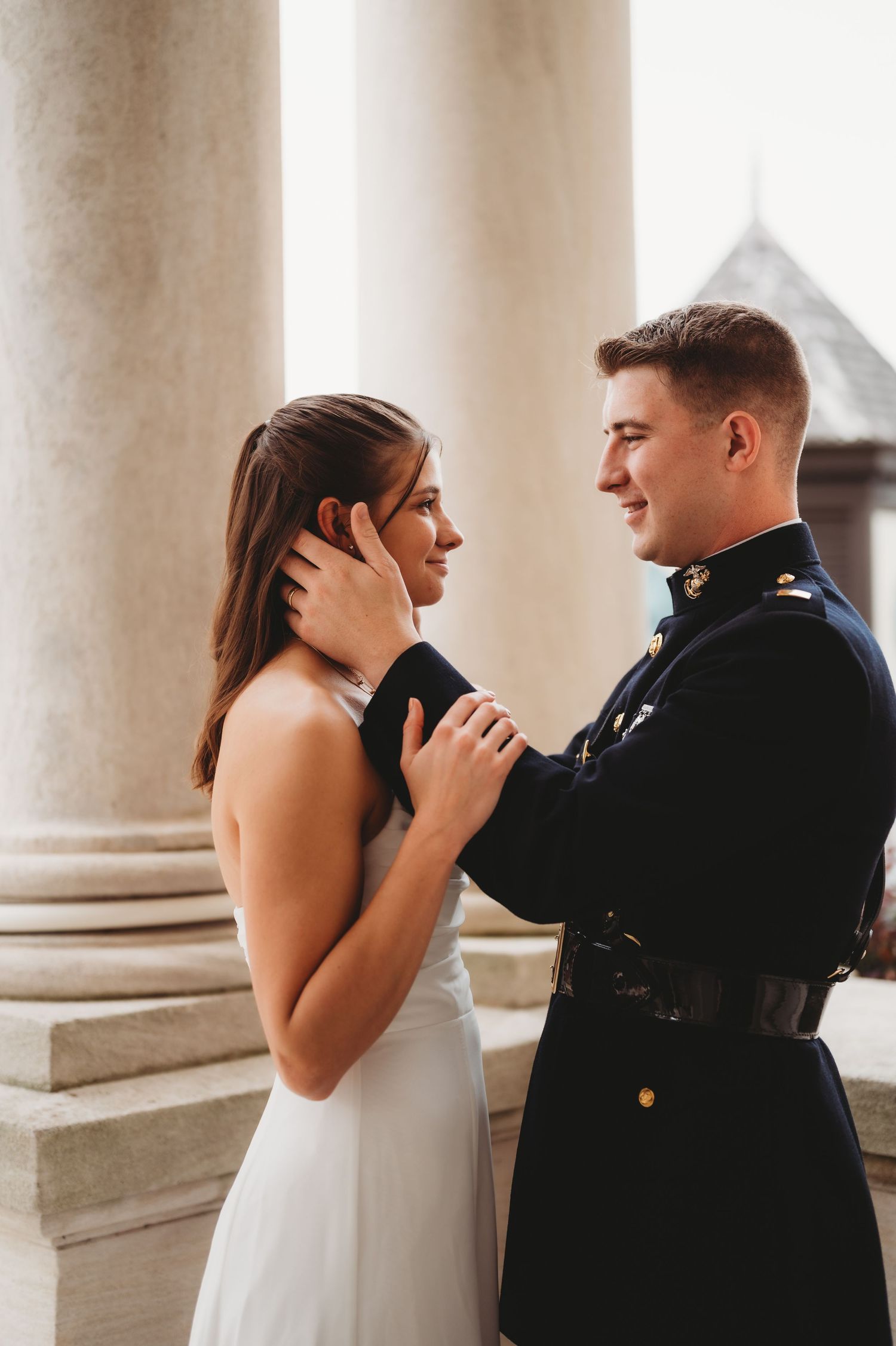 A military groom in dress uniform tenderly holds a bride's face near grand columns.