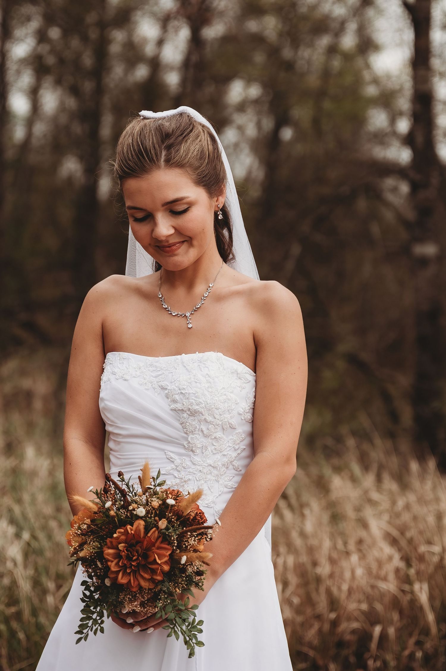 Bride in strapless white gown and veil holds autumn bouquet of orange dahlias, smiling softly outdoors.