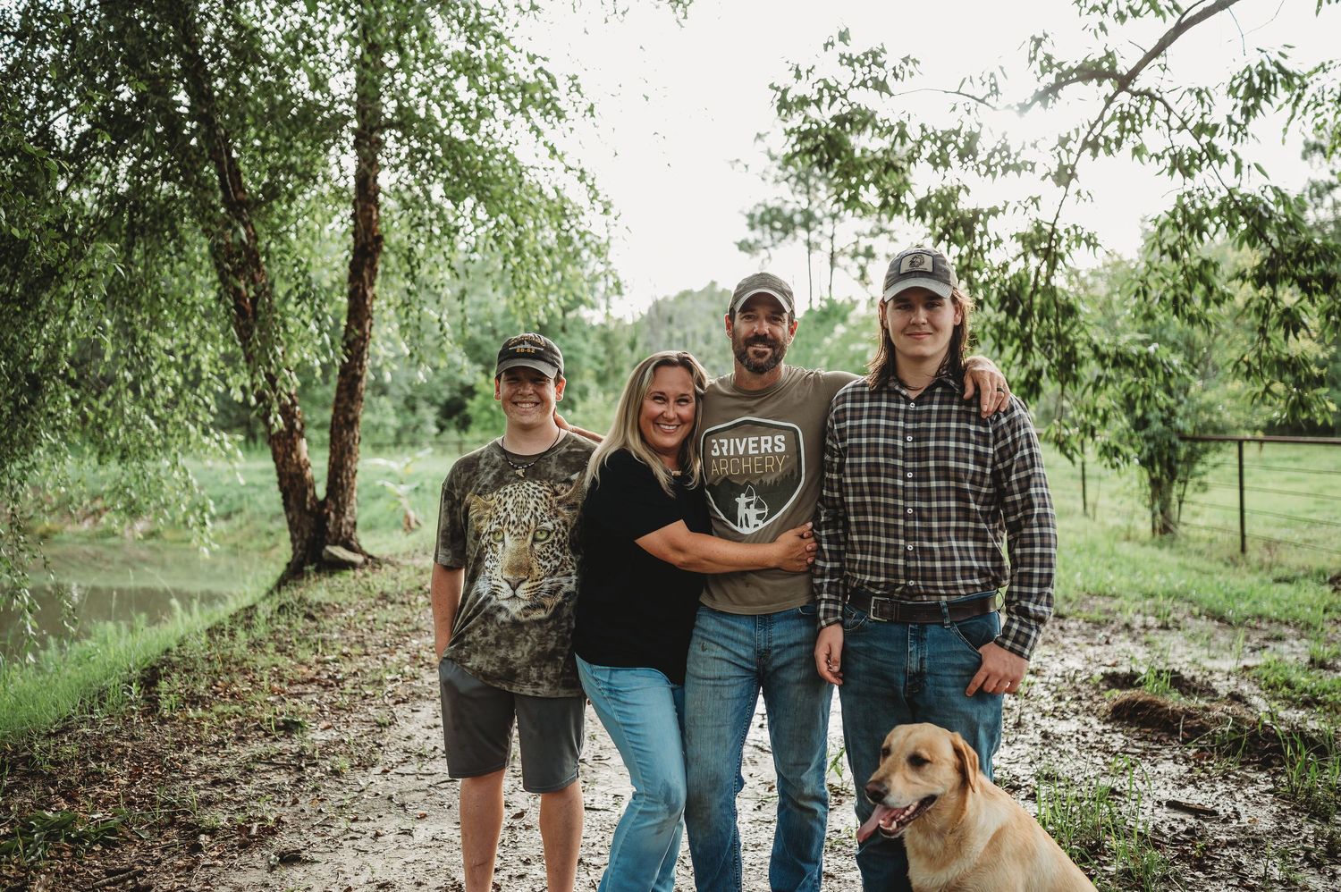 Family of four posing outdoors on a rural path with a yellow Labrador dog, surrounded by lush green trees.