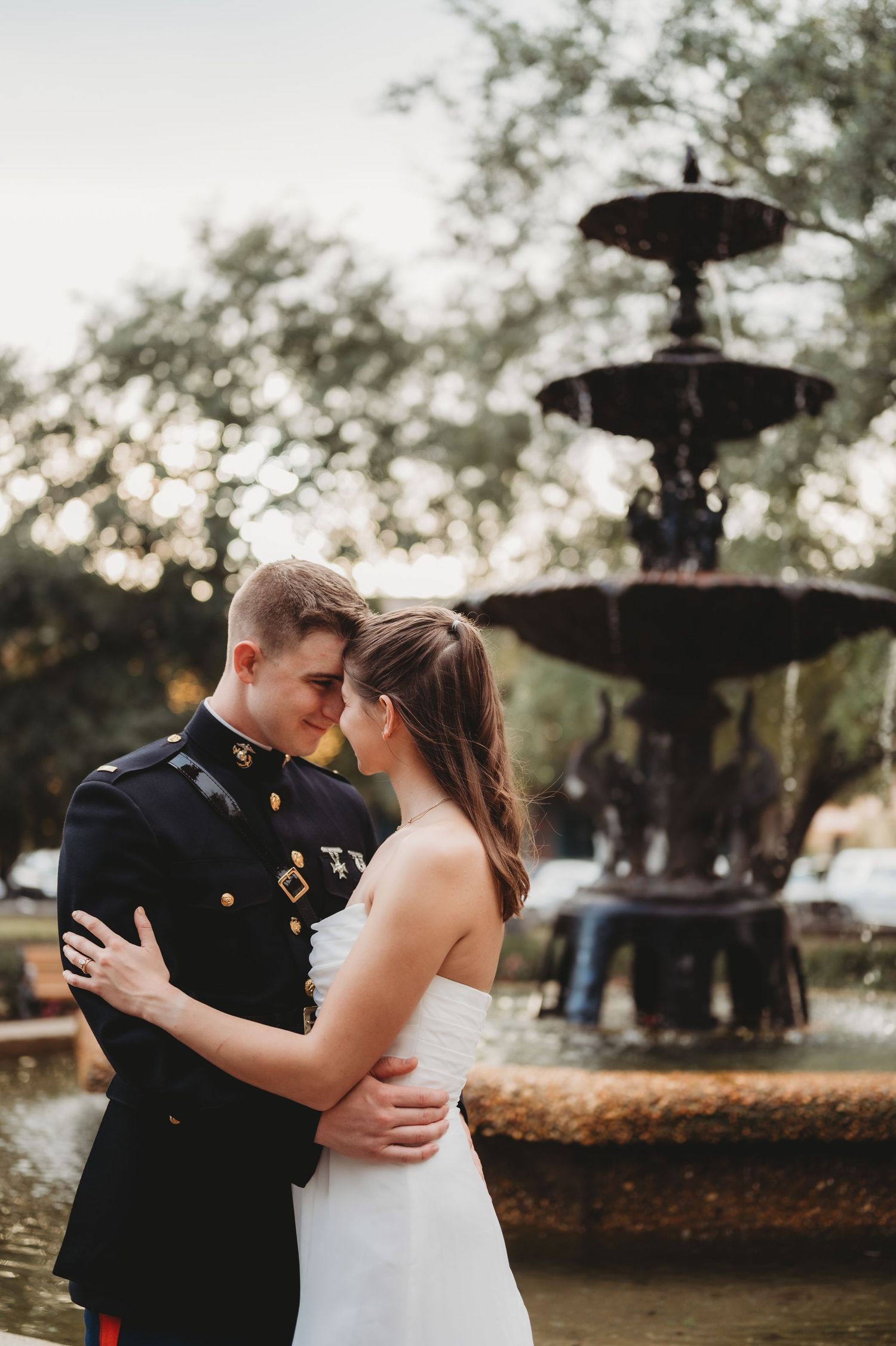 Bride and groom in military dress uniform embrace romantically in front of an ornate fountain.