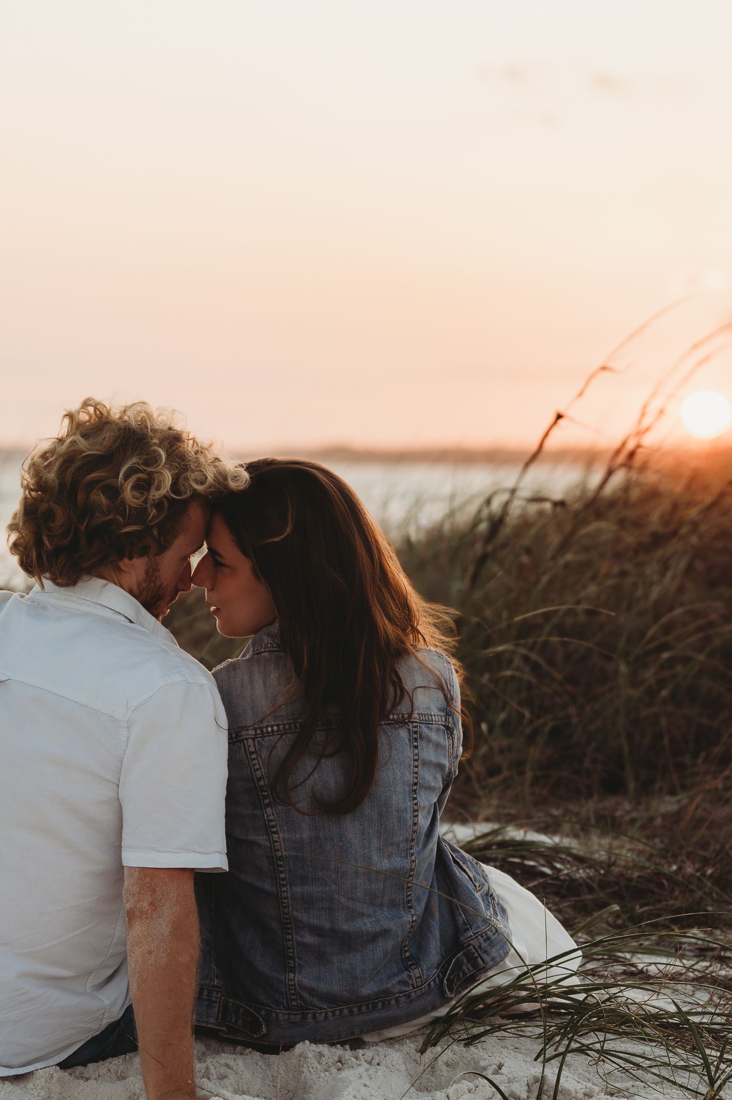 Couple sitting together on beach dunes at sunset, sharing a romantic moment with tall grass and golden sky.