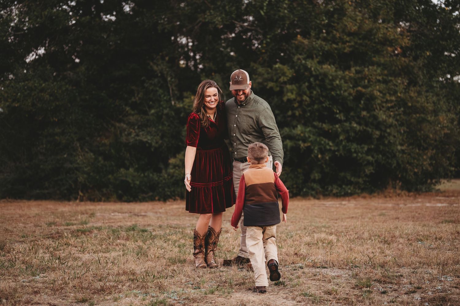 Family of three smiling and walking together in an open field surrounded by trees during autumn.