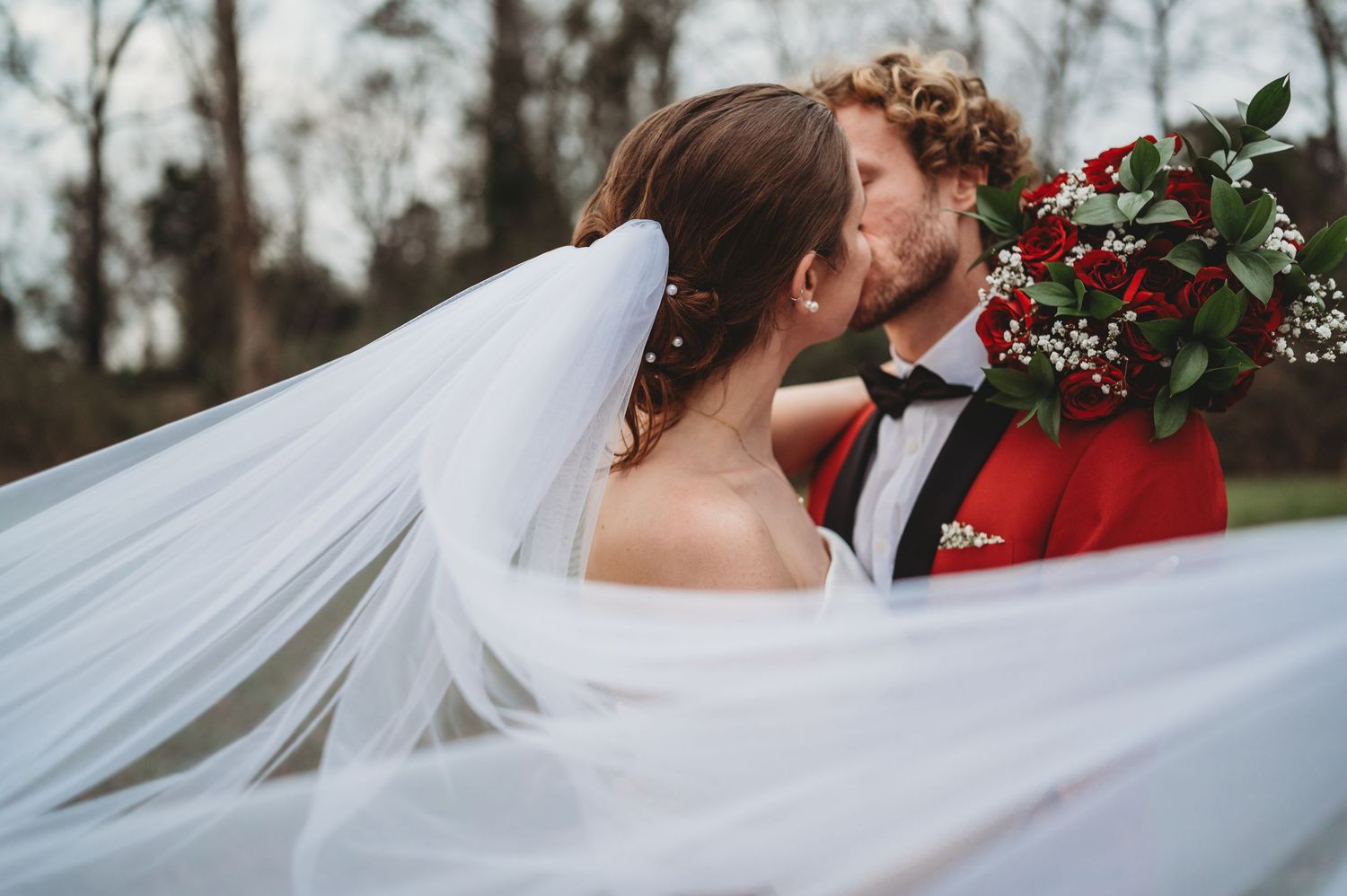 Bride and groom sharing a kiss outdoors, with flowing white veil and red floral bouquet on their wedding day.