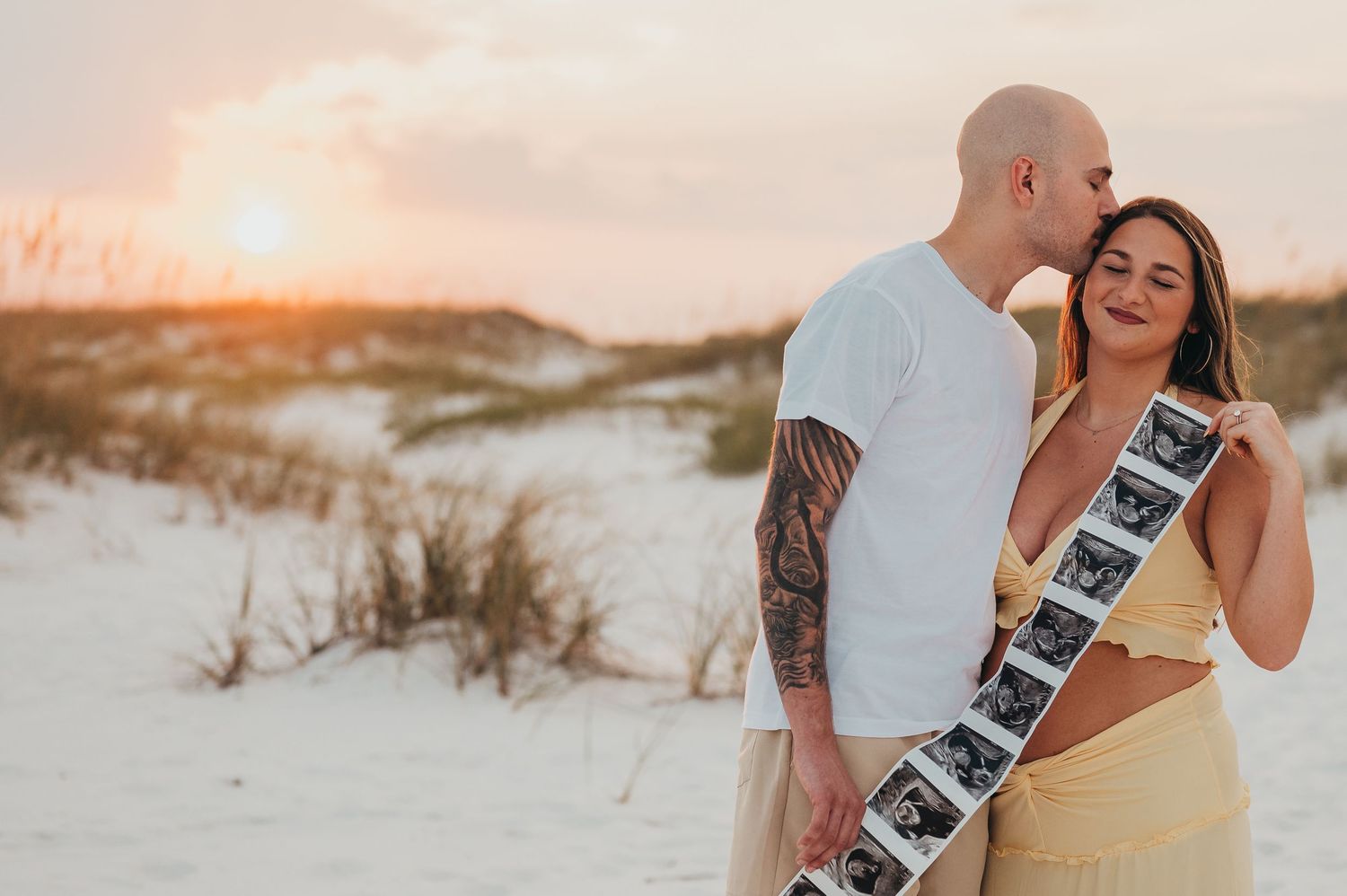 Couple embracing on sandy beach at sunset, holding ultrasound photos, announcing pregnancy in romantic coastal setting.
