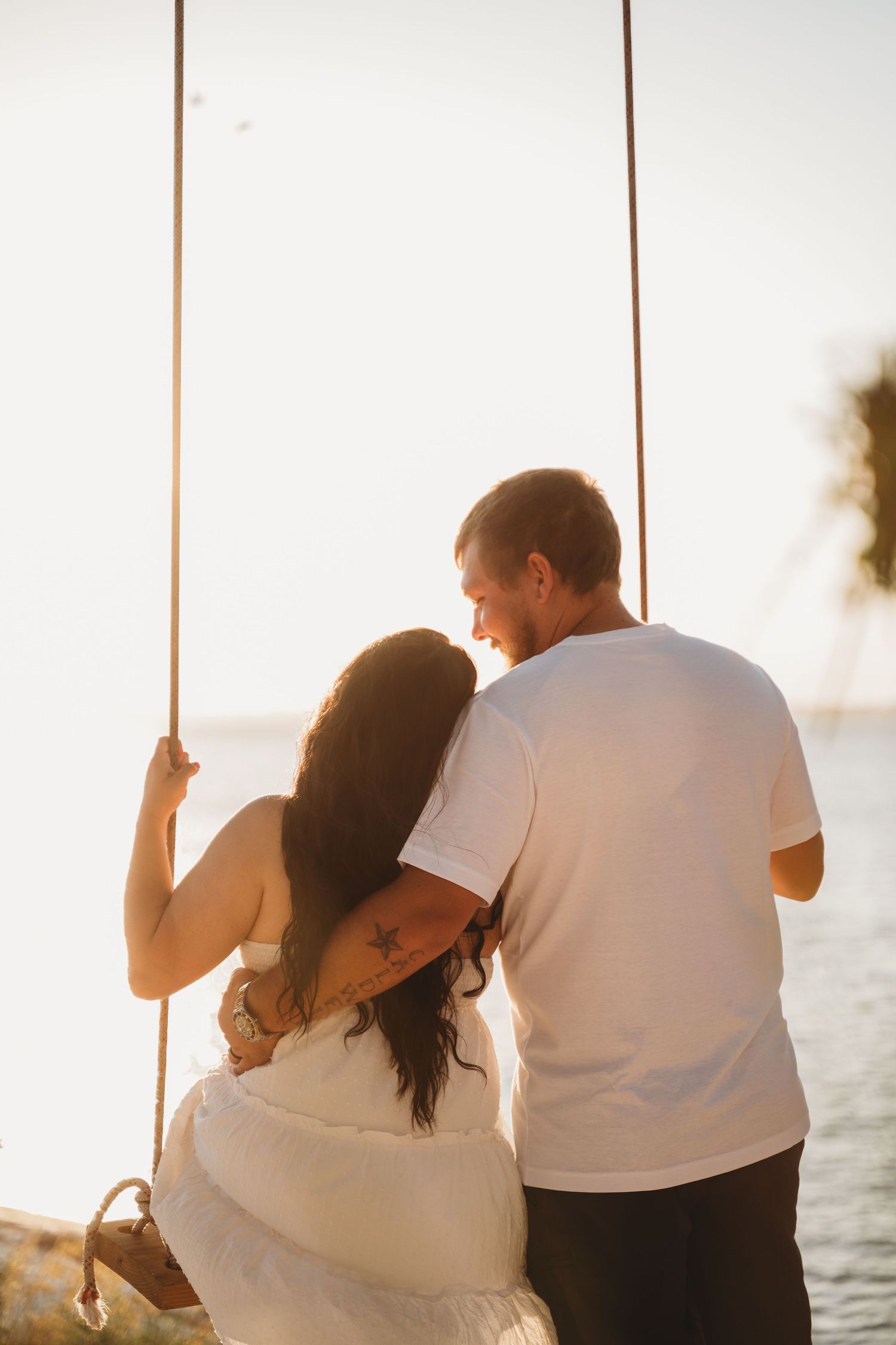 A couple shares a romantic moment on a swing overlooking the ocean at sunset.