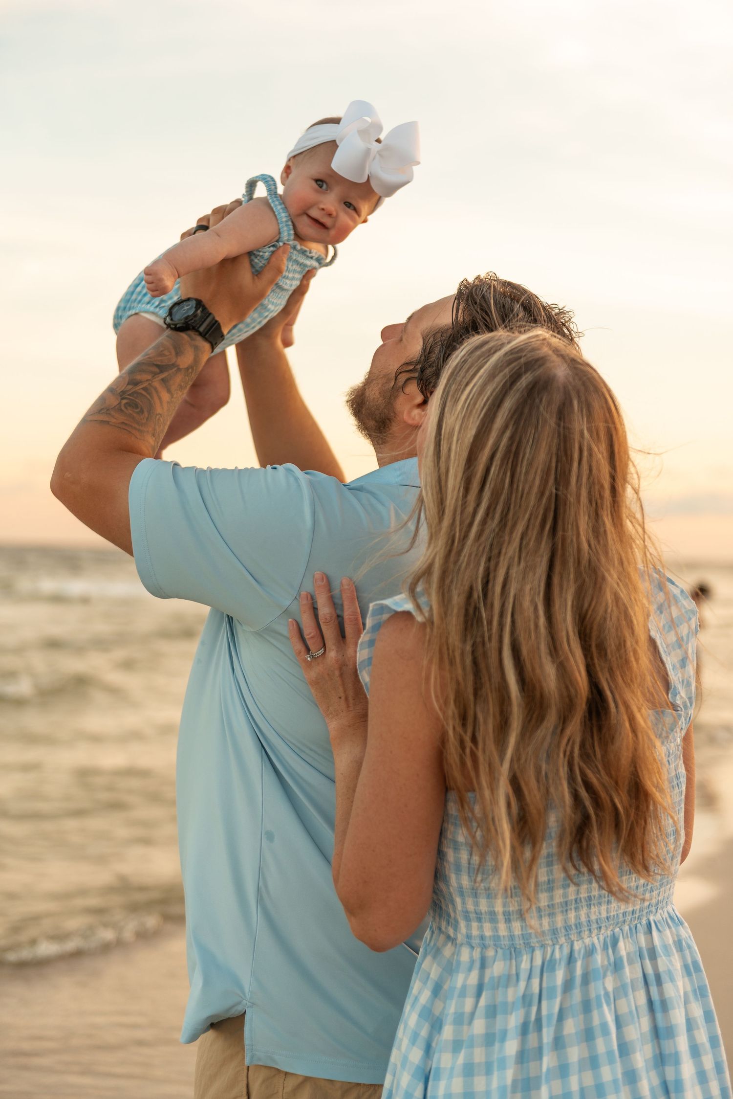 Parents lifting their baby girl with a white bow at a beach during a warm sunset.