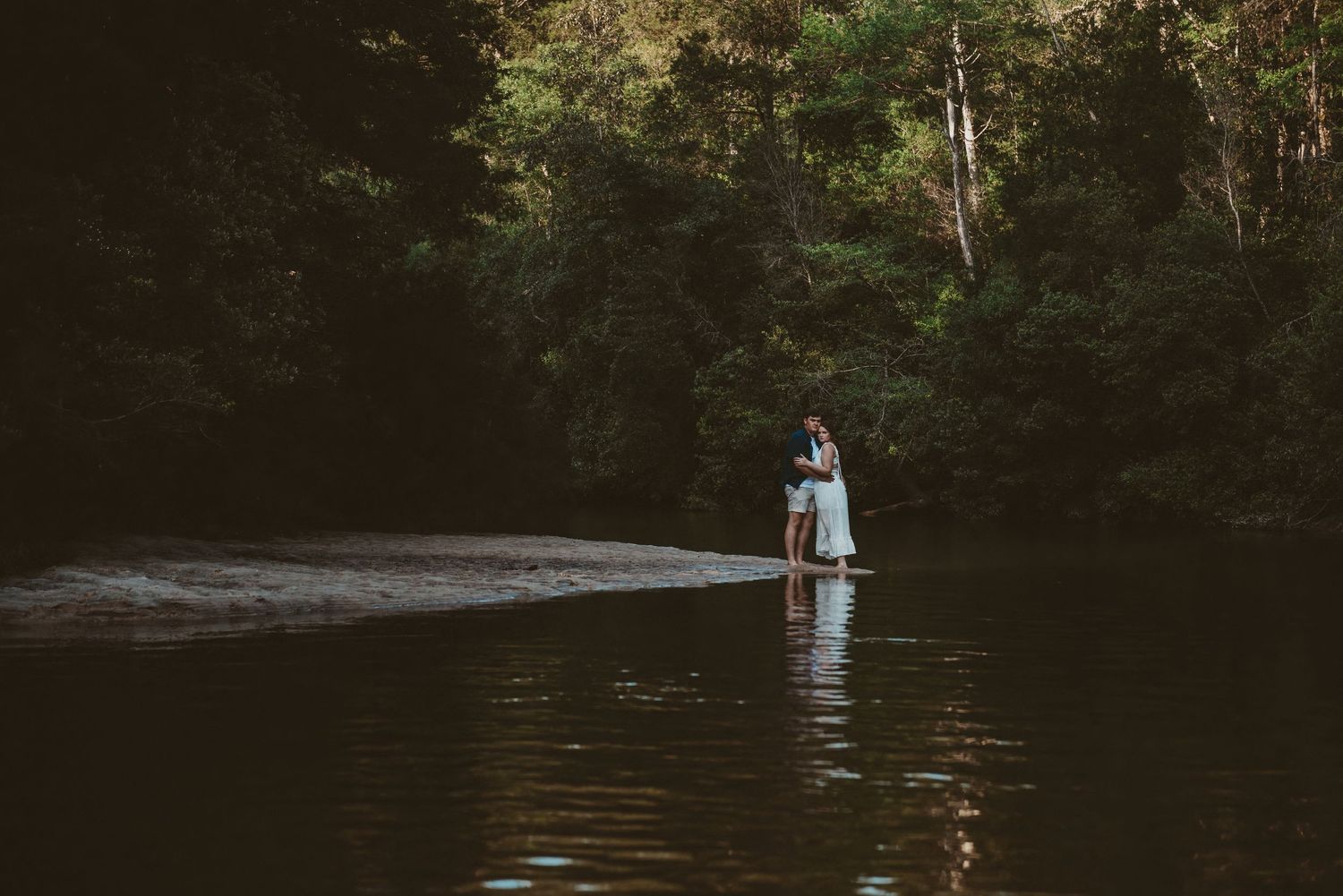 Couple standing together in shallow river water surrounded by dense green forest trees.
