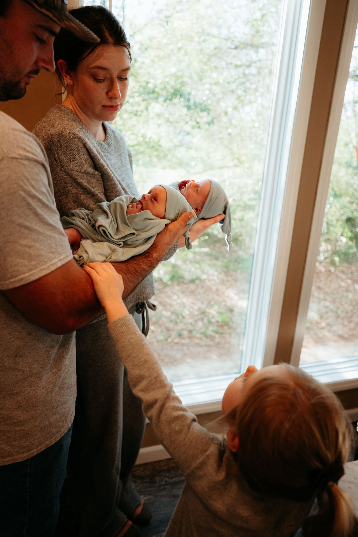 Parents hold newborn baby near window as curious young child reaches up to touch the infant.