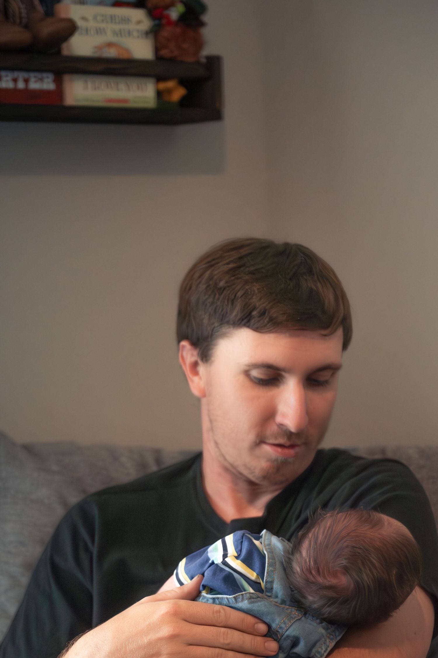 New father in black shirt gently holding his sleeping newborn baby on a couch.