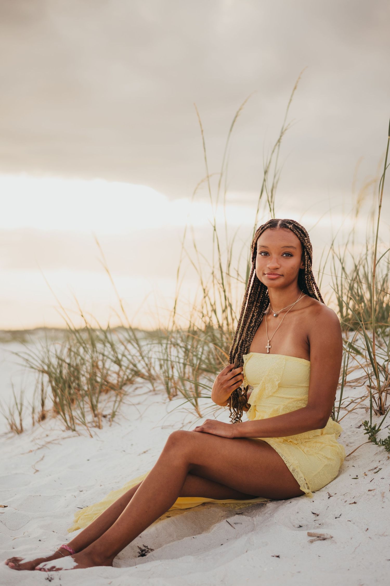 Teen girl in yellow strapless dress with braids sitting on sand dunes surrounded by beach grass.