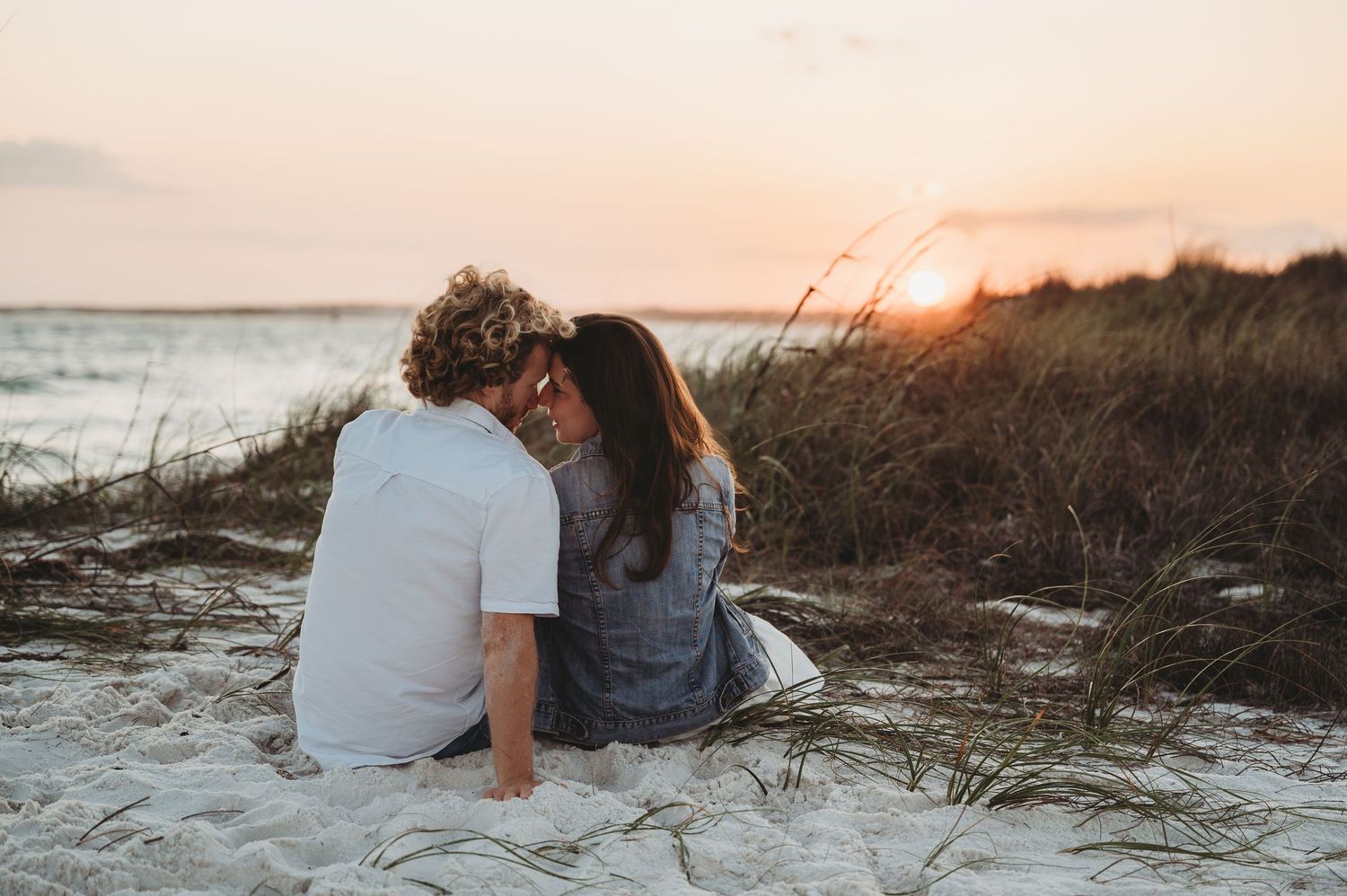 Couple sharing a romantic sunset kiss on a sandy beach surrounded by coastal grass and golden evening light.
