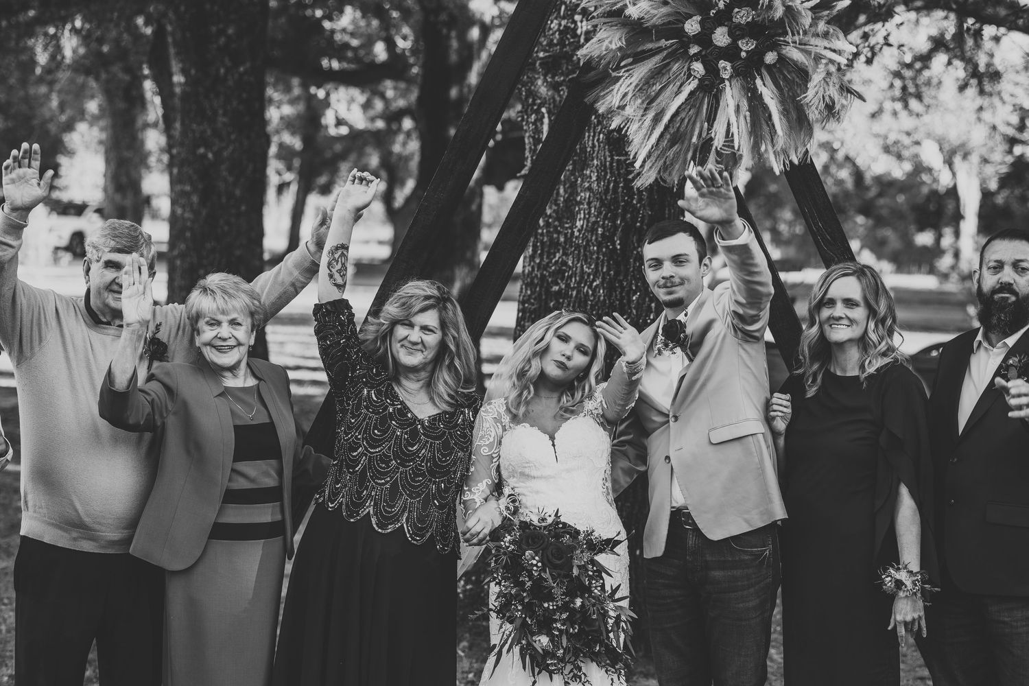 Black and white photo of joyful wedding party celebrating with raised arms under a teepee arch.
