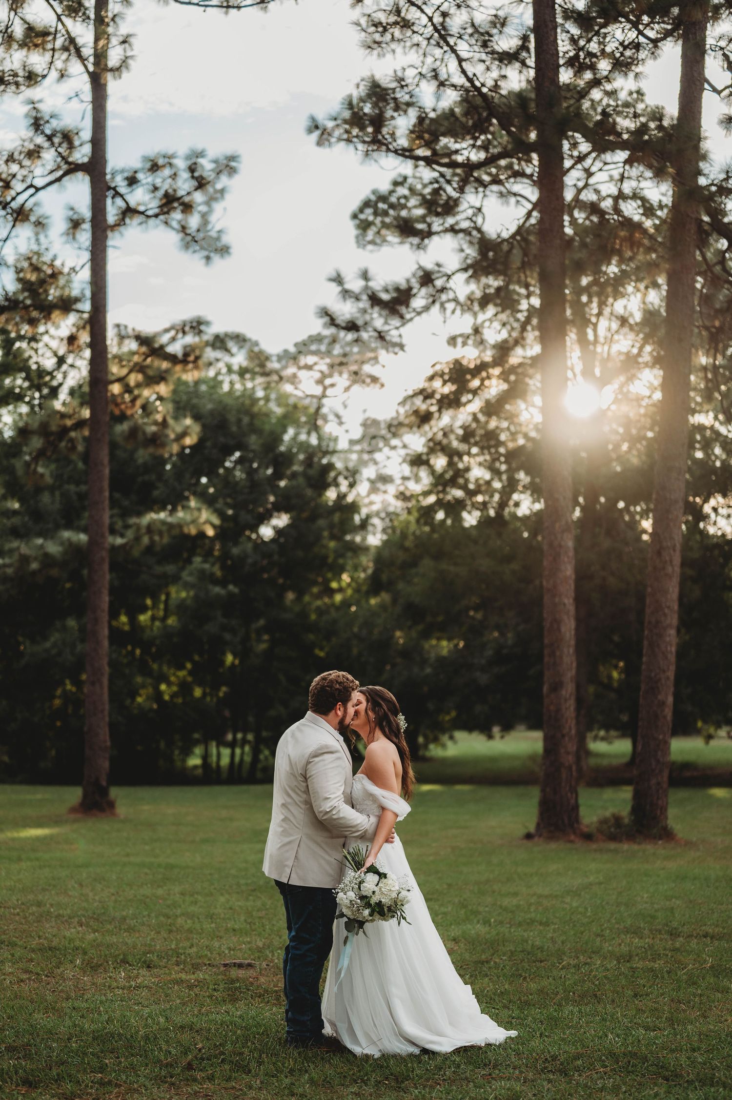 Bride and groom kissing at golden hour surrounded by tall pine trees in a lush green field.