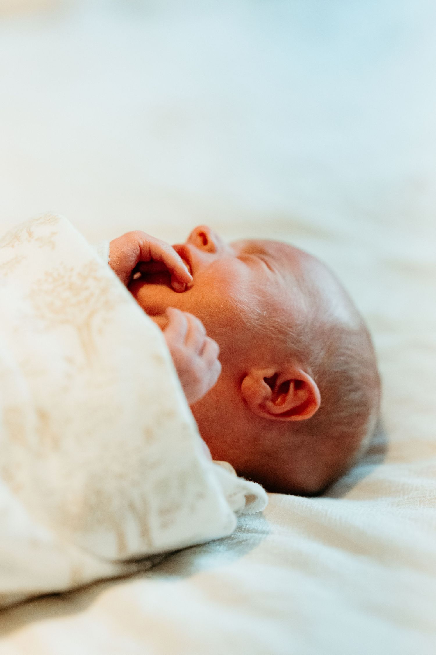 Newborn baby swaddled in white blanket lying on soft surface with tiny hand raised.