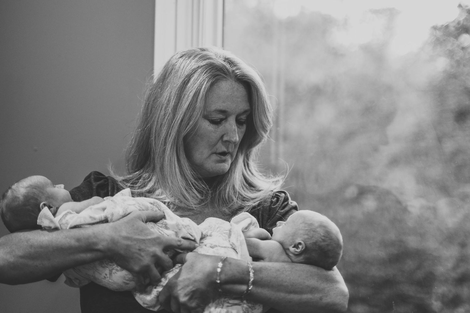 Blonde mother gazing lovingly at twin newborn babies held in both arms, black and white.