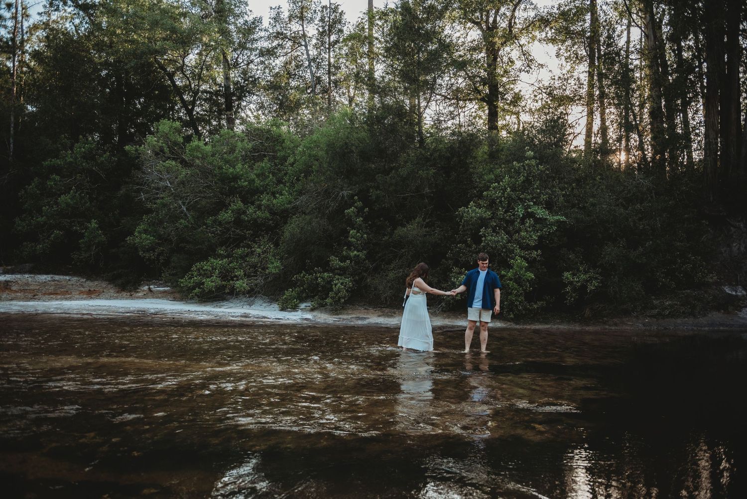 Couple holding hands while wading through a shallow river near a small waterfall in a forest.