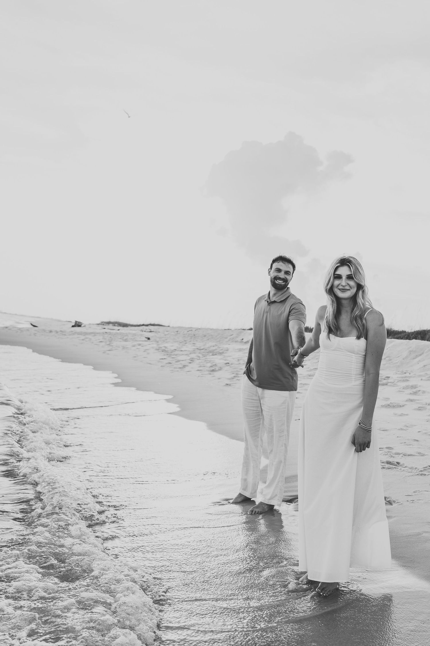 Black and white photo of a couple holding hands and walking along the shoreline at the beach.