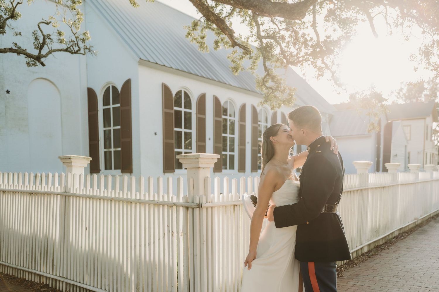 Bride and groom share a romantic kiss outside a charming blue church with white picket fence at golden hour.