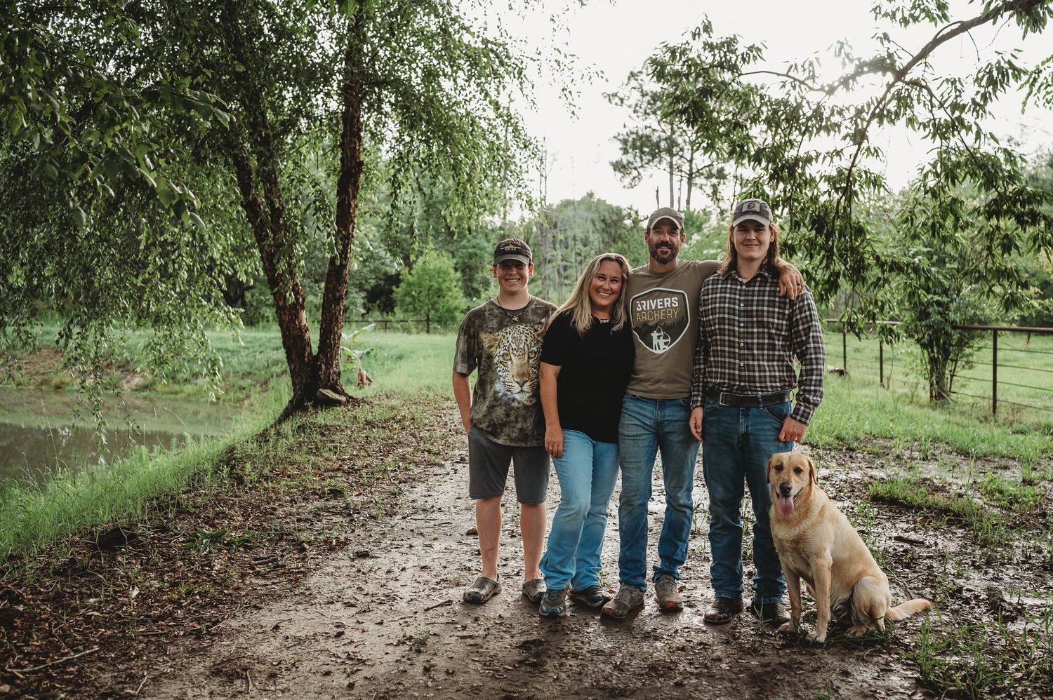 Four people and a golden retriever dog posing together on a muddy rural path surrounded by lush green trees.