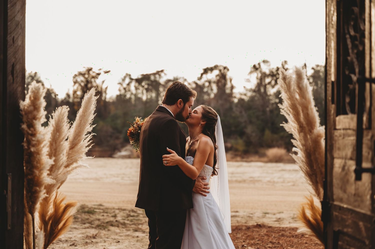 Bride and groom sharing a romantic kiss at barn doorway, framed by tall pampas grass, with golden field backdrop.