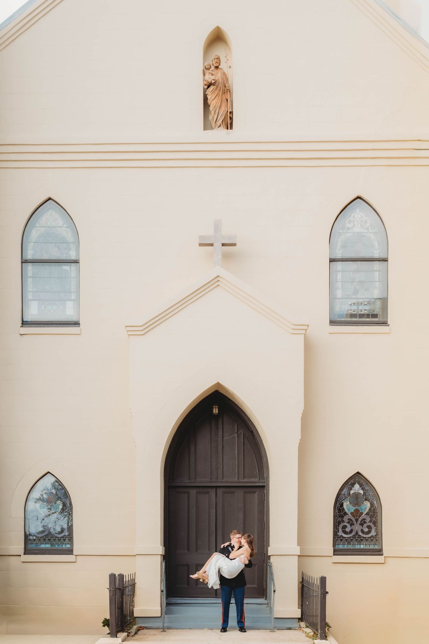 Groom lifting bride in wedding attire outside a white church with arched windows and a cross.