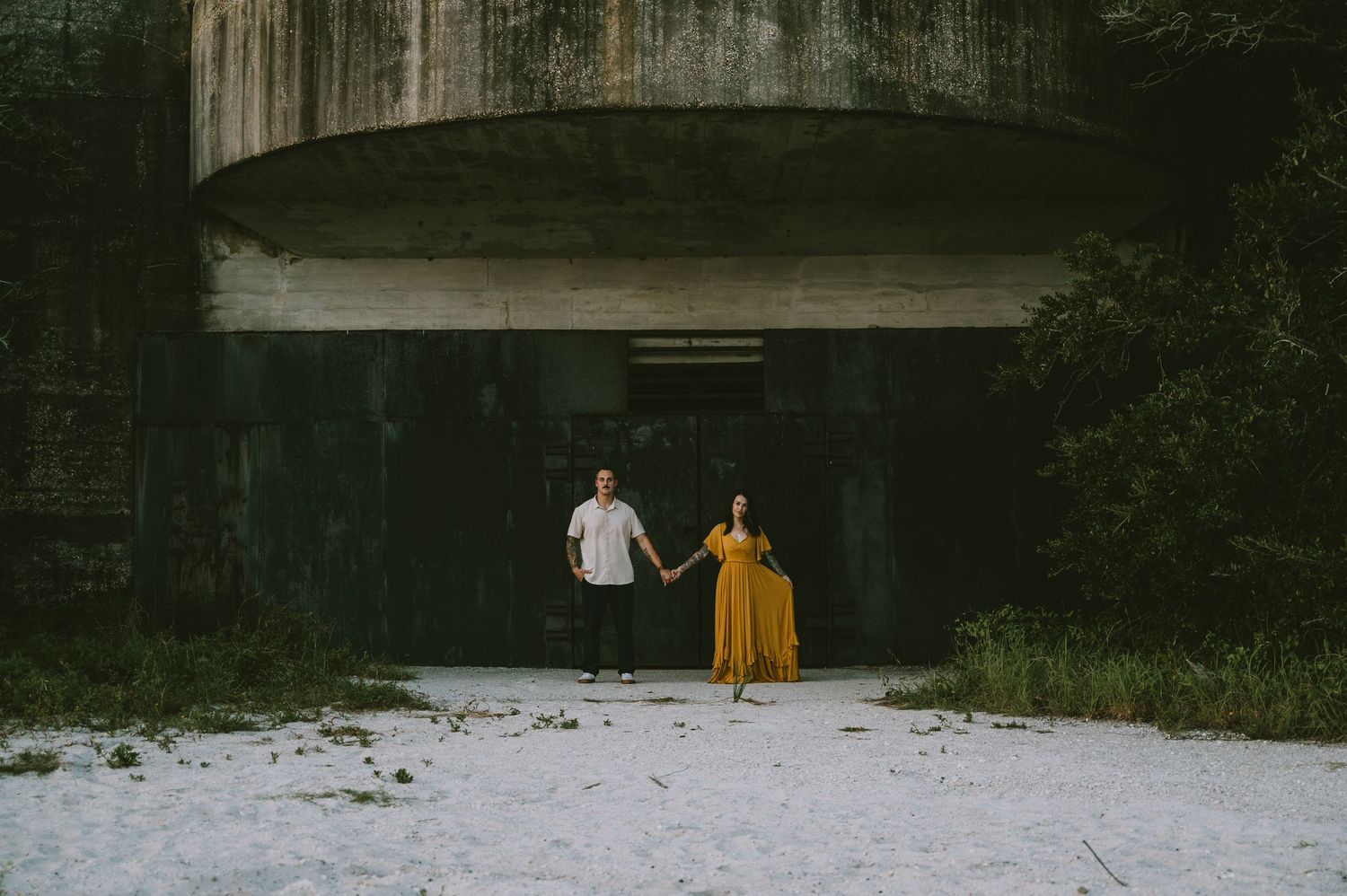 Couple holding hands in front of a concrete structure surrounded by trees.