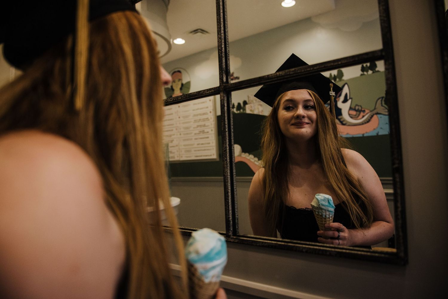 Smiling graduate in cap and gown holding blue ice cream cone while looking at her reflection in a mirror.