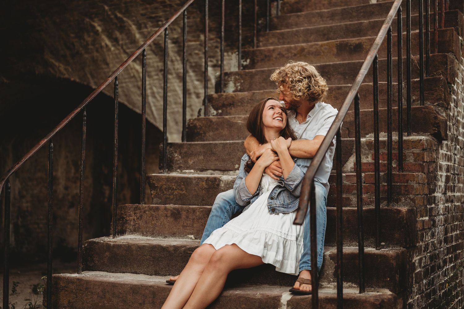 Romantic couple embracing on rustic brick staircase, woman in white dress, man in denim jacket kissing her cheek.