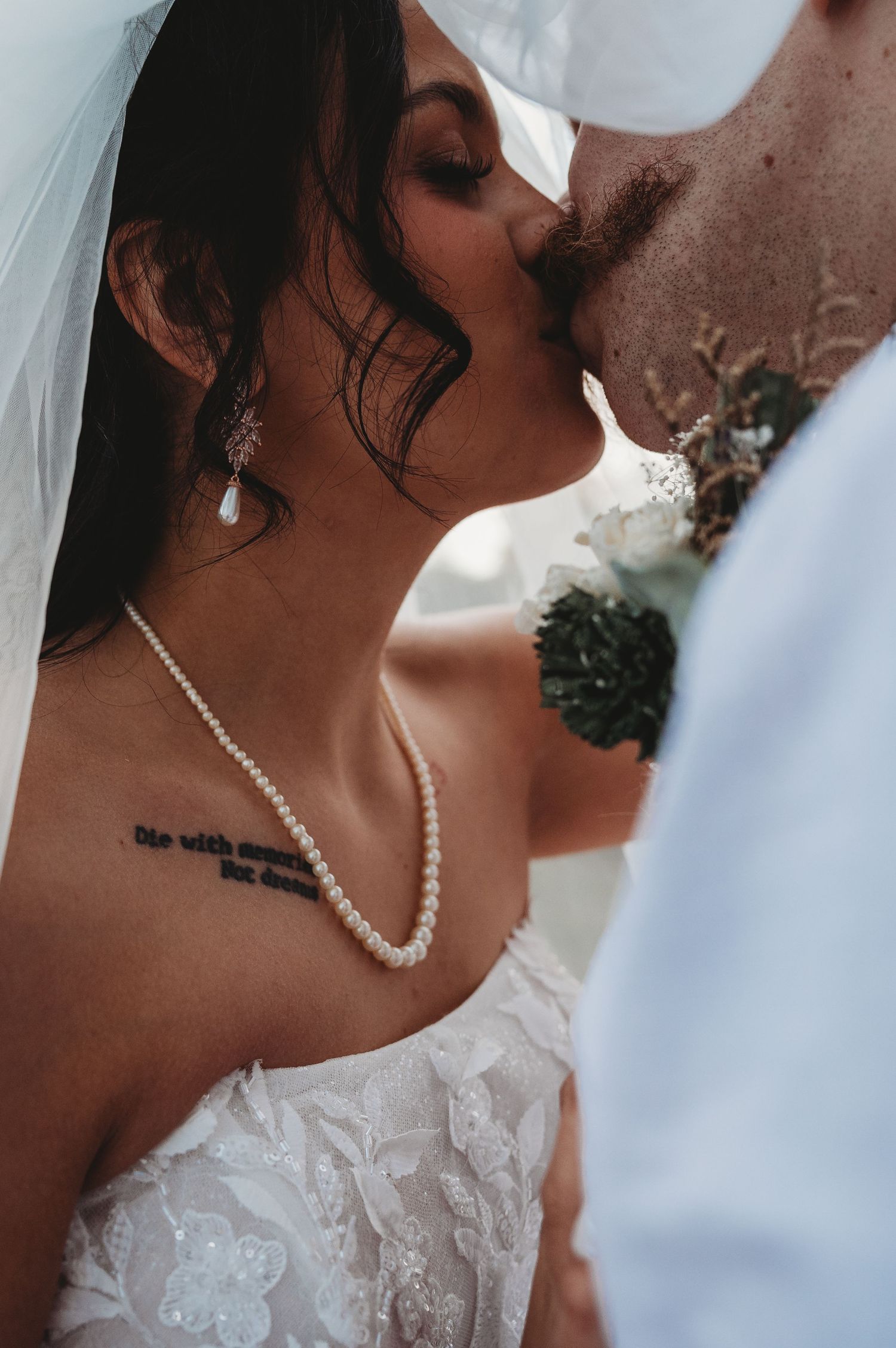 Bride and groom sharing a romantic kiss, bride wearing pearl necklace and lace gown with veil on wedding day.