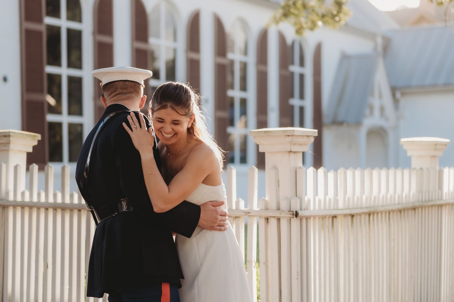 Military groom in dress uniform embraces smiling bride in white gown near white picket fence at outdoor wedding venue.