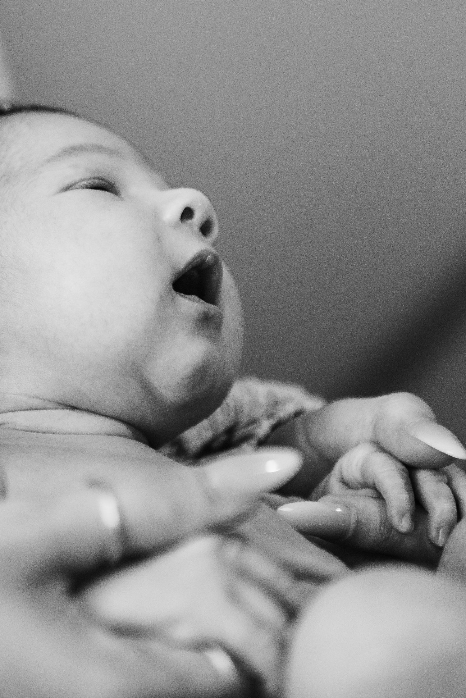 Black and white close-up of newborn baby yawning while adult gently holds tiny hands.