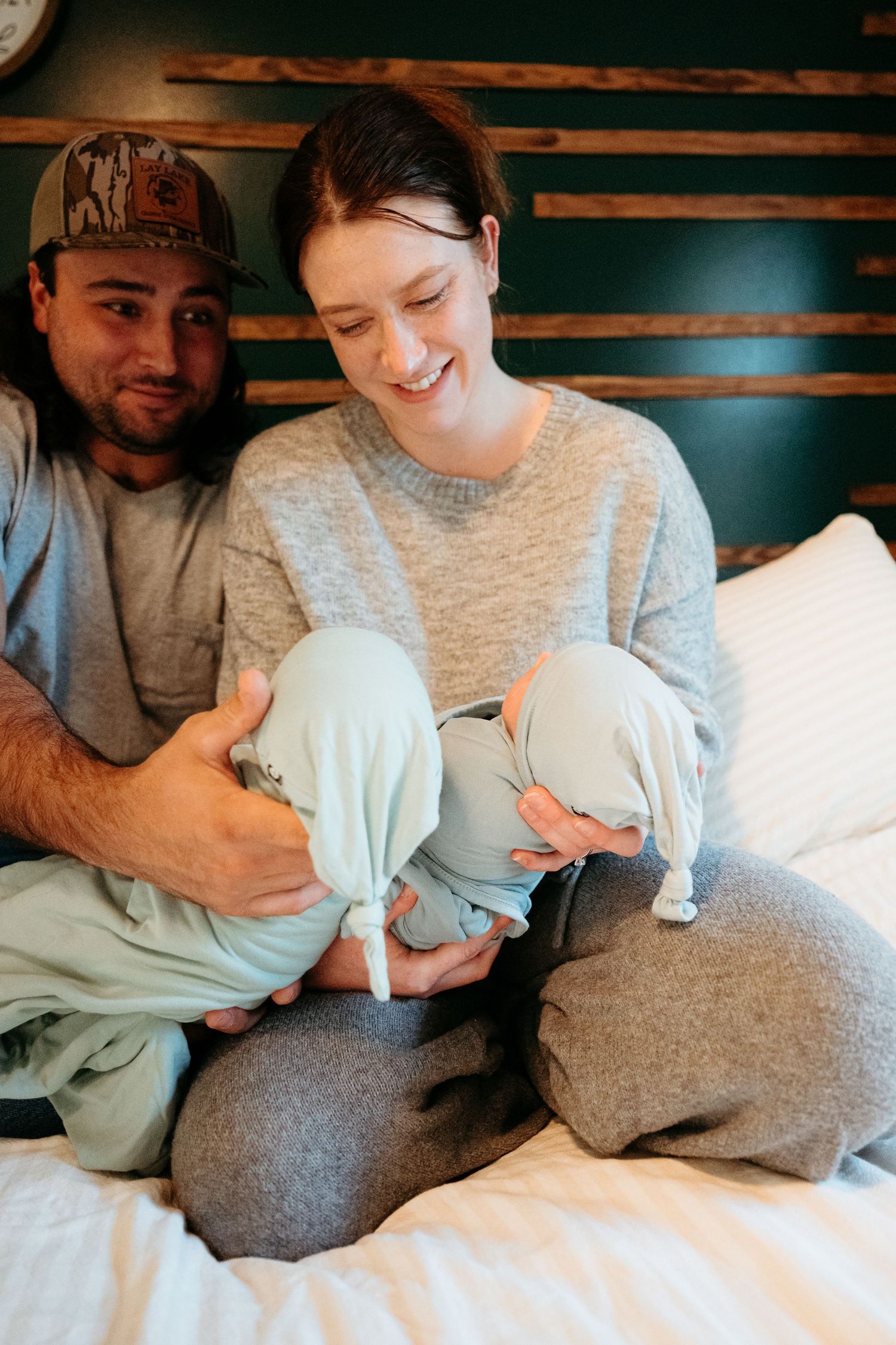 Smiling parents sitting on a white bed holding their twin newborn babies in light blue swaddles.