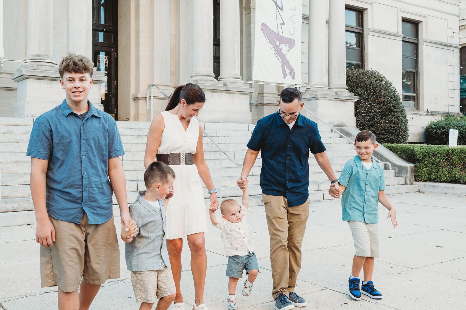 A family of six walks together hand-in-hand outside a grand classical building.