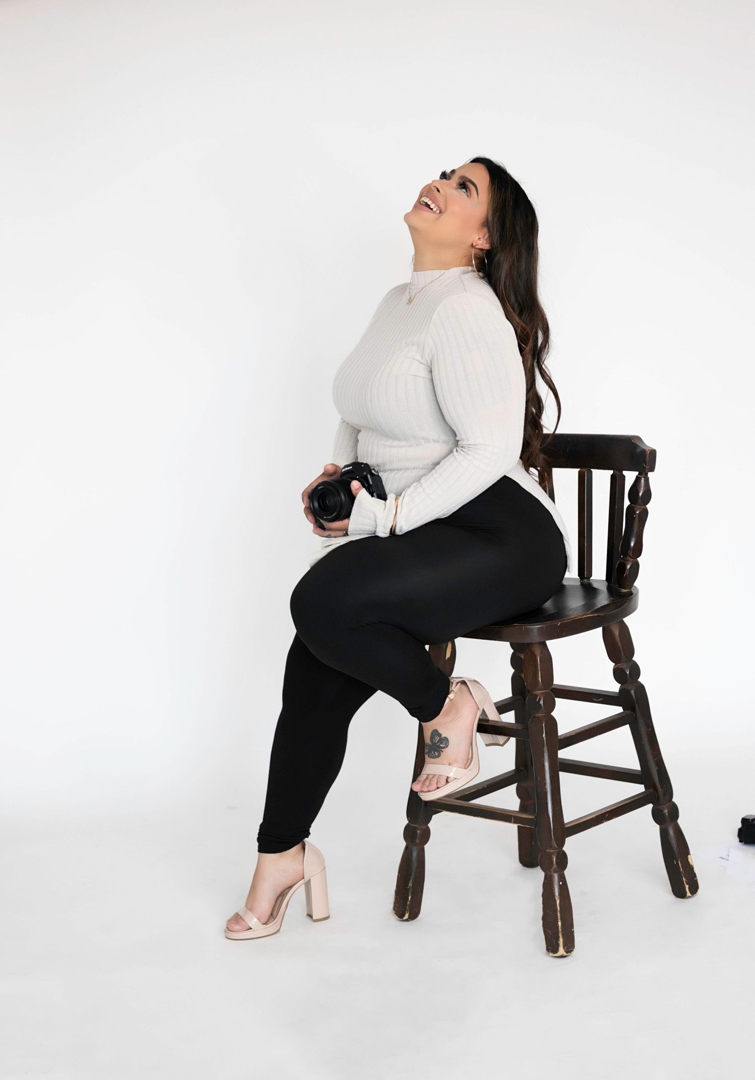 Woman in white sweater and black leggings sits on wooden stool holding camera, looking up against white background.