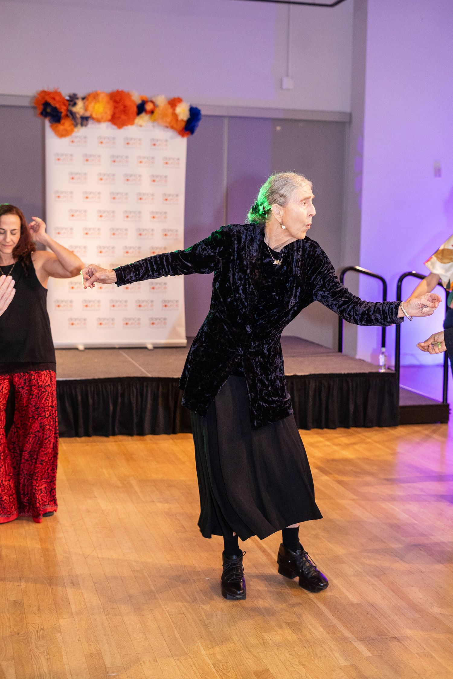 A woman in black velvet dress dances gracefully on a wooden floor at a formal event with a branded backdrop.