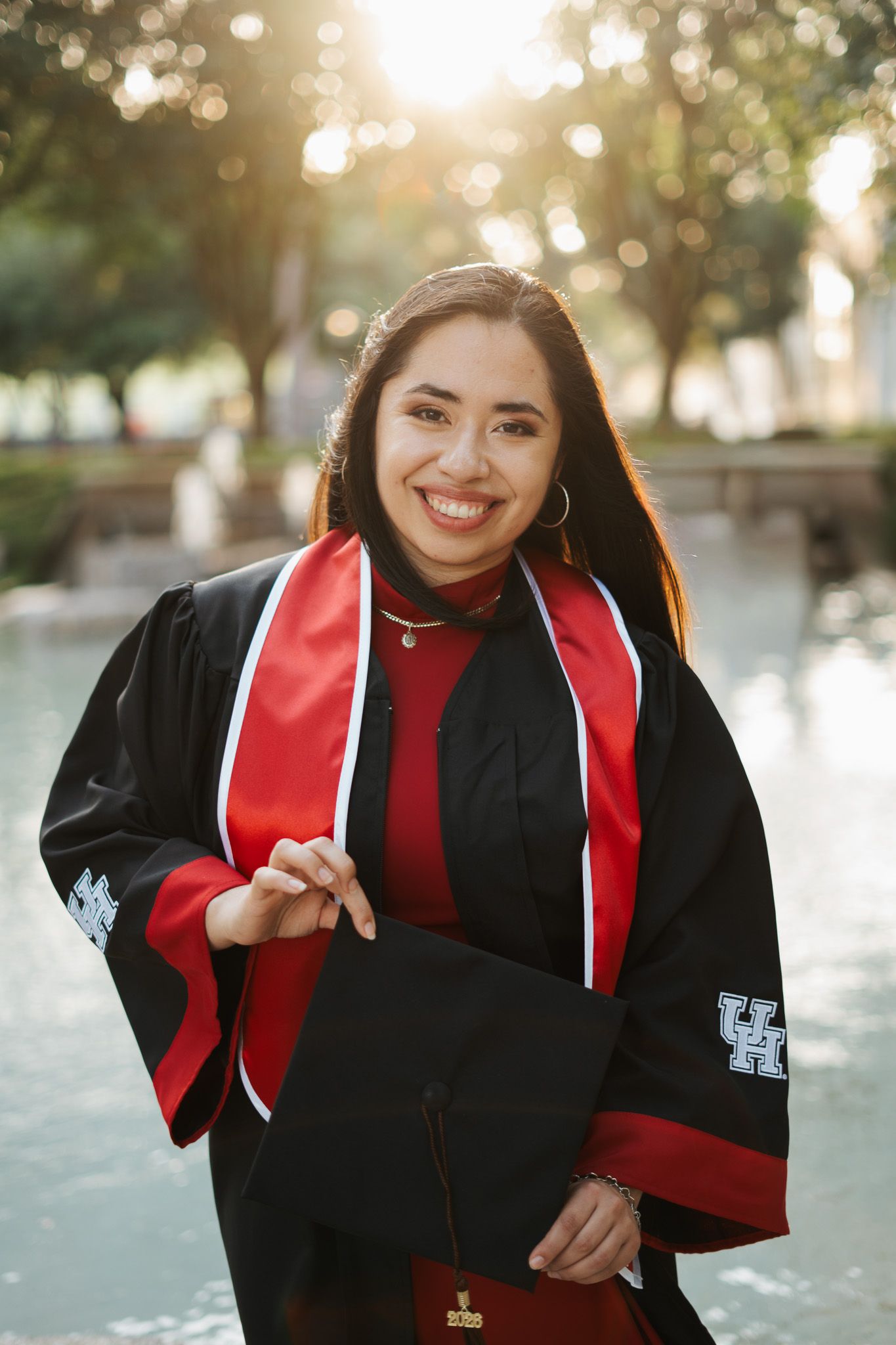Smiling female graduate in black gown with red stole holding cap at University of Houston graduation ceremony outdoors.