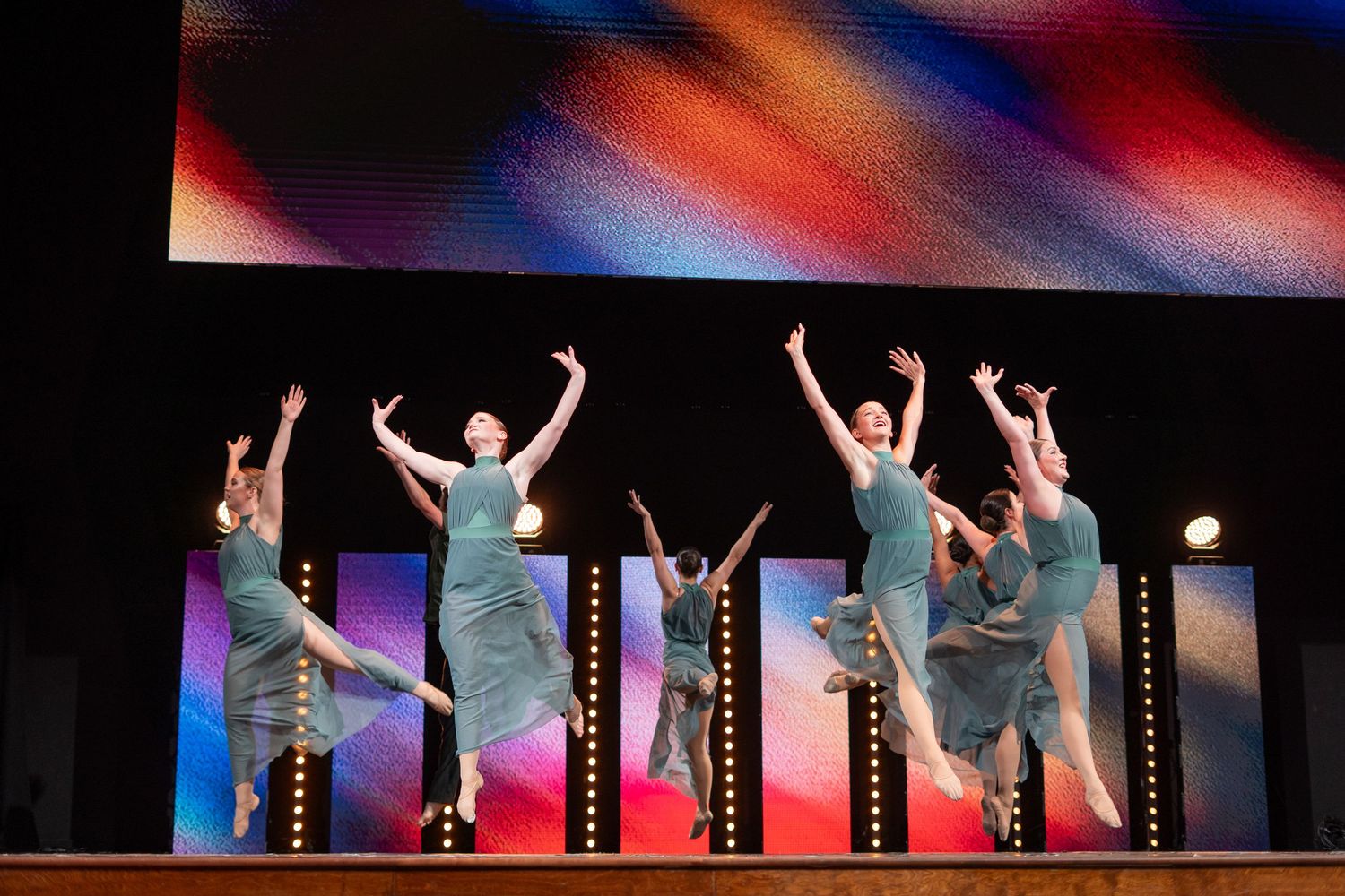 Five dancers in teal costumes leap gracefully on a colorful lit stage during a synchronized dance performance.