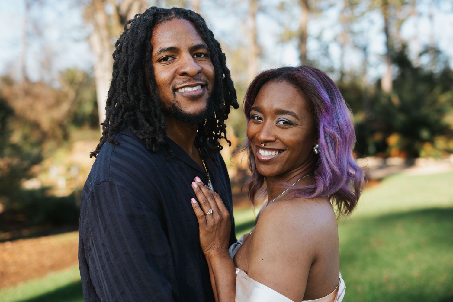 Happy couple smiling outdoors, woman with purple hair showing engagement ring, man with dreadlocks, in a park setting.