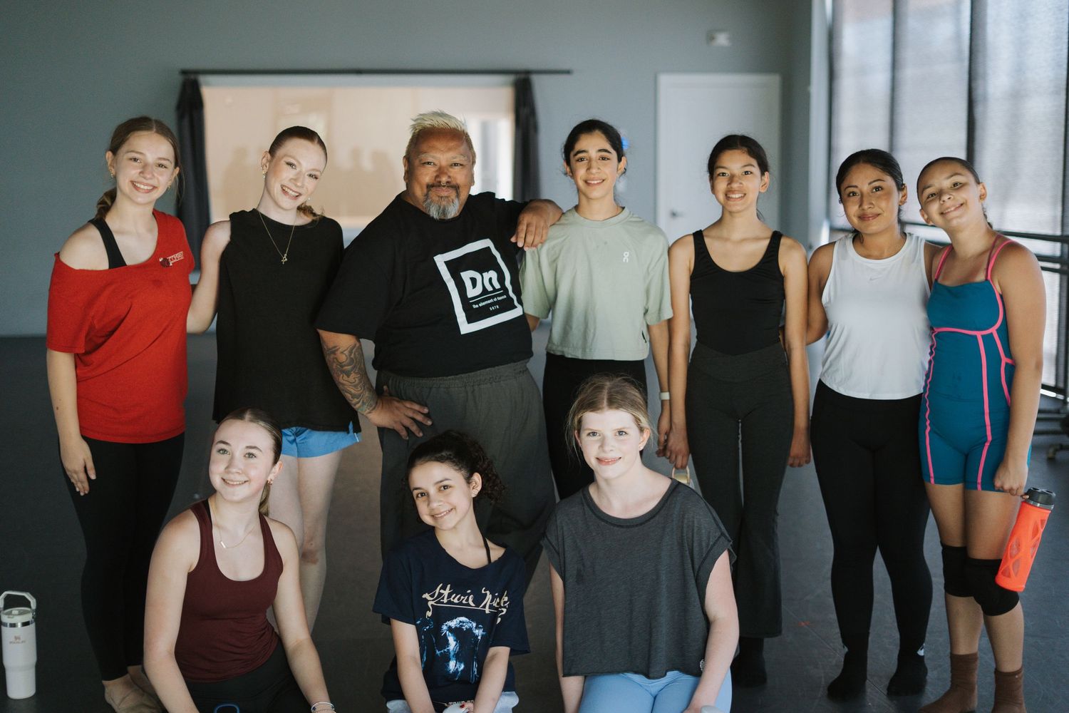 A dance instructor poses with a group of young female students in a bright studio setting.