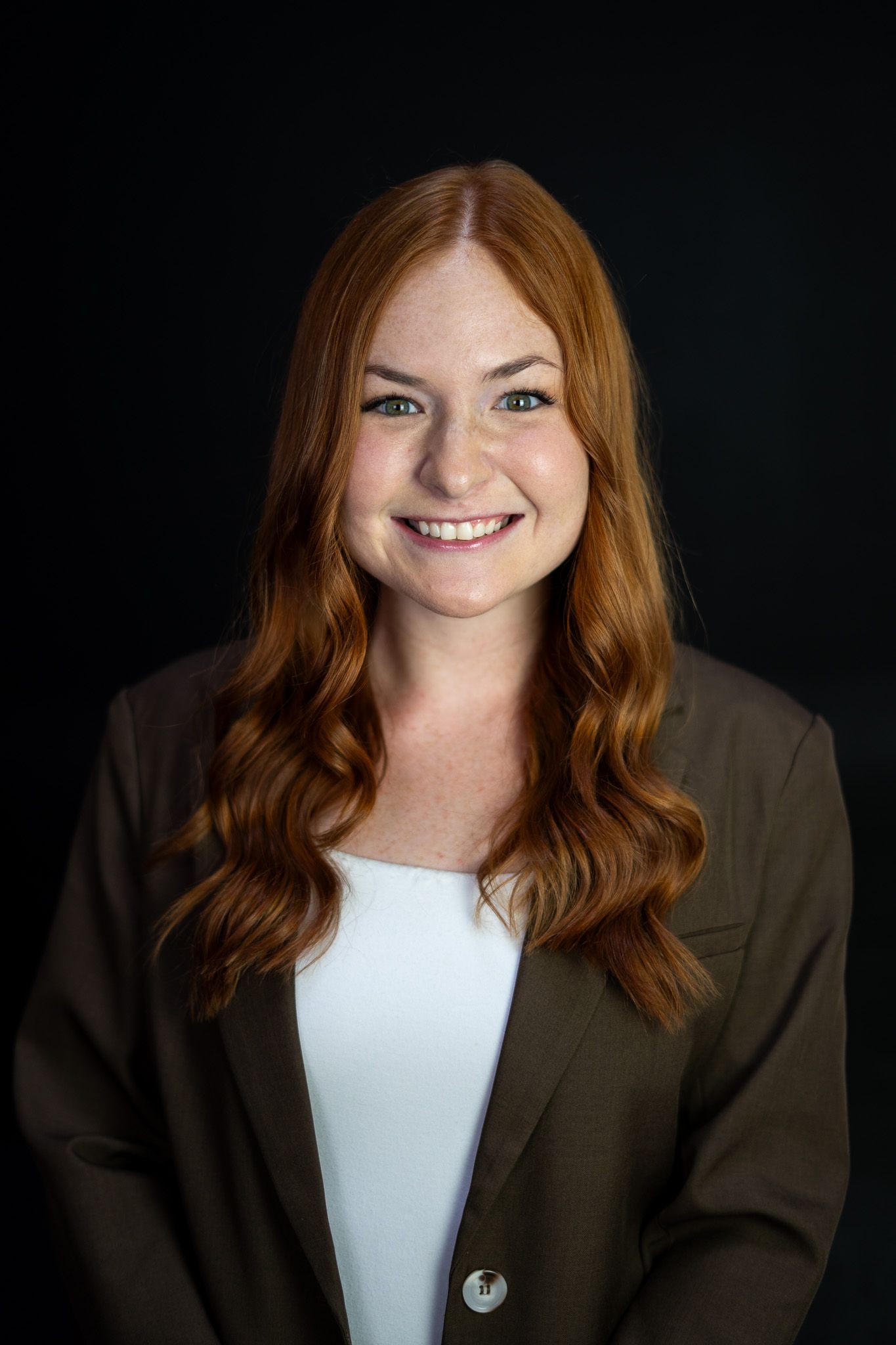 A smiling young woman with long red hair wearing a dark blazer poses for a professional headshot on a black background.