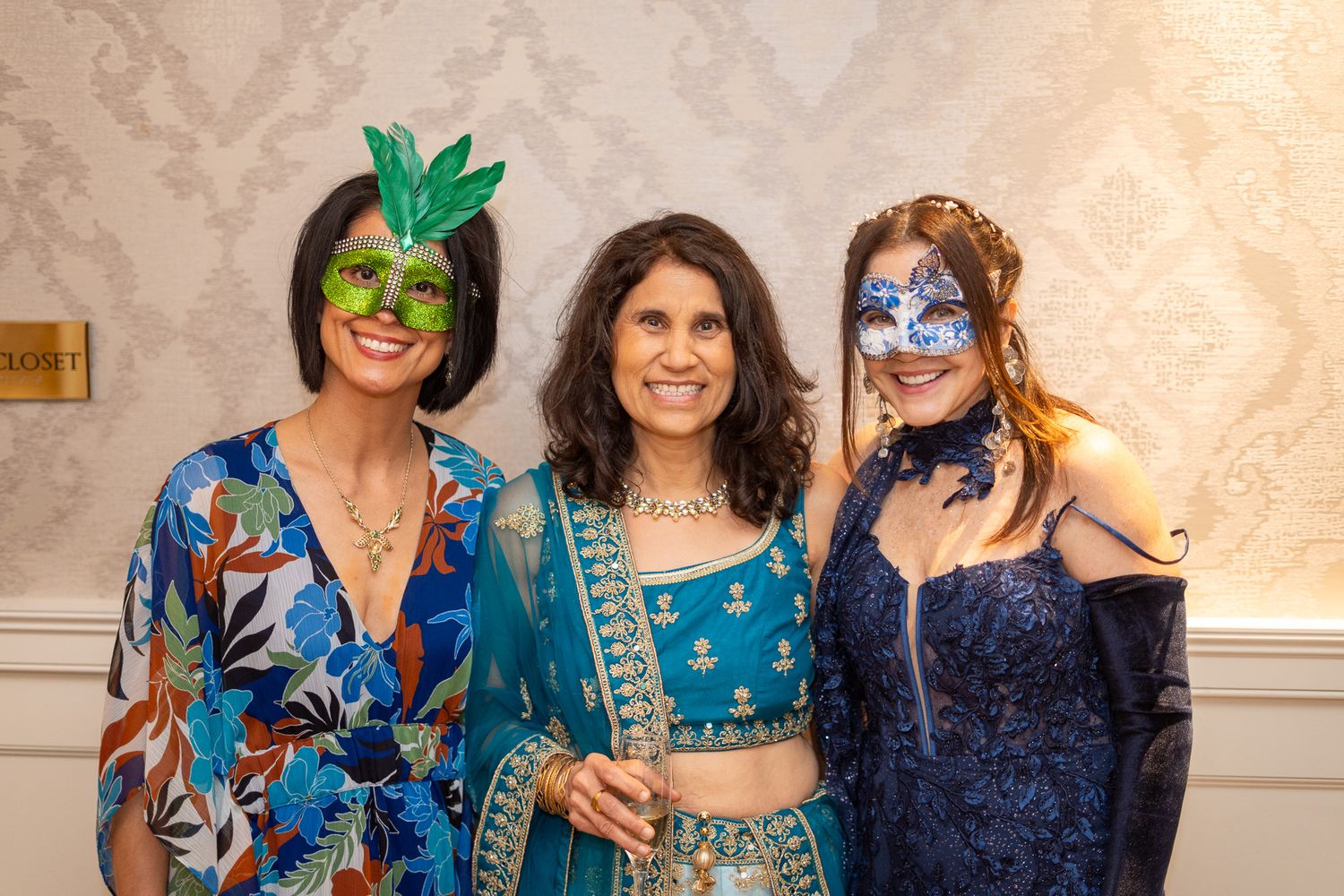 Three women in masquerade masks pose together at a formal masquerade gala event.