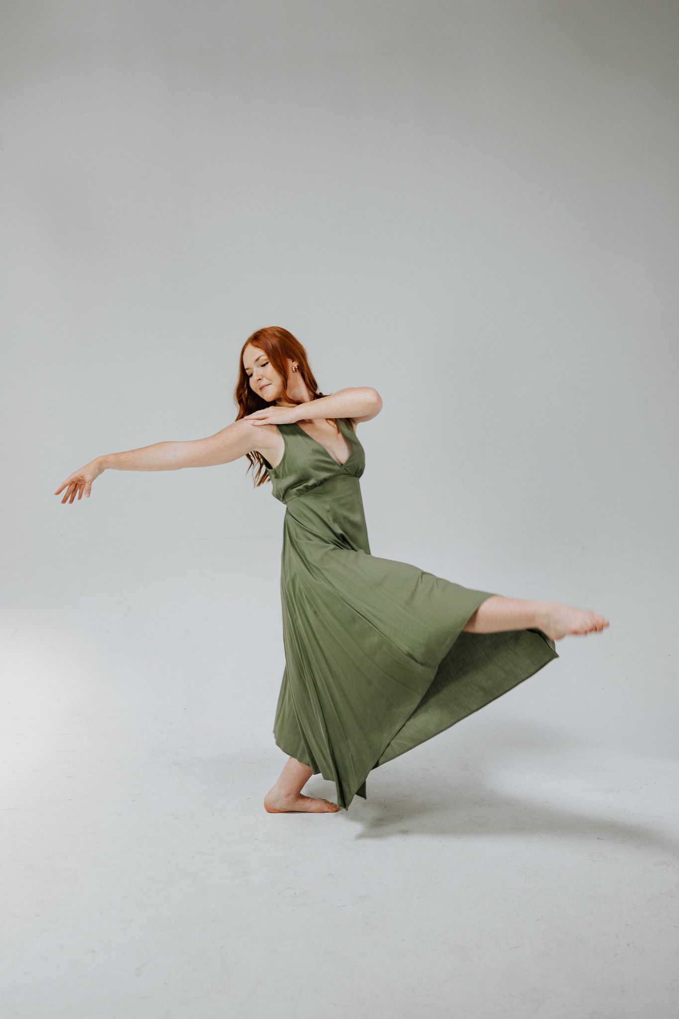 A barefoot woman in a flowing green dress poses gracefully mid-dance against a light gray background.
