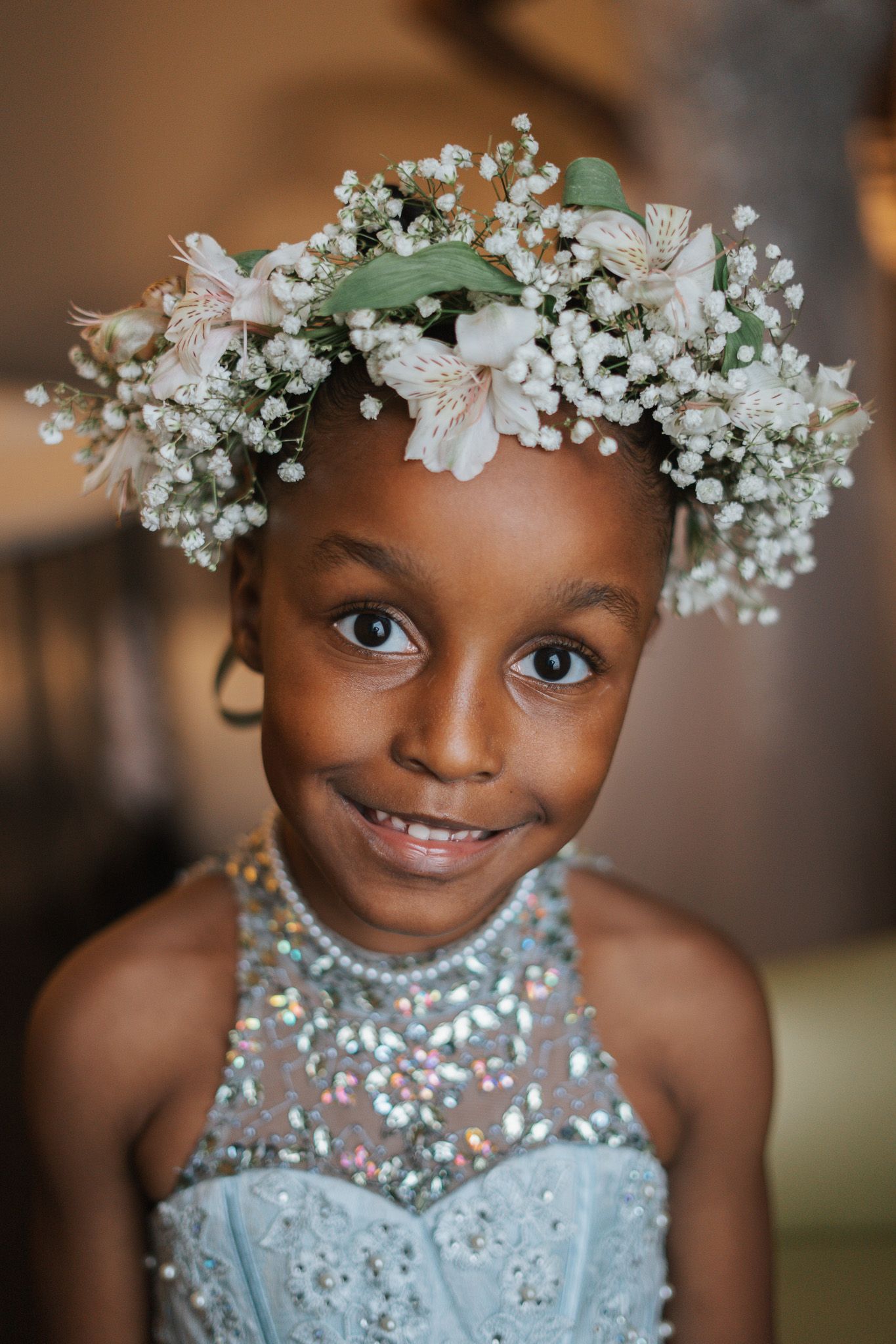 Young flower girl smiling in beaded blue dress wearing floral crown of white baby's breath and pink blooms.