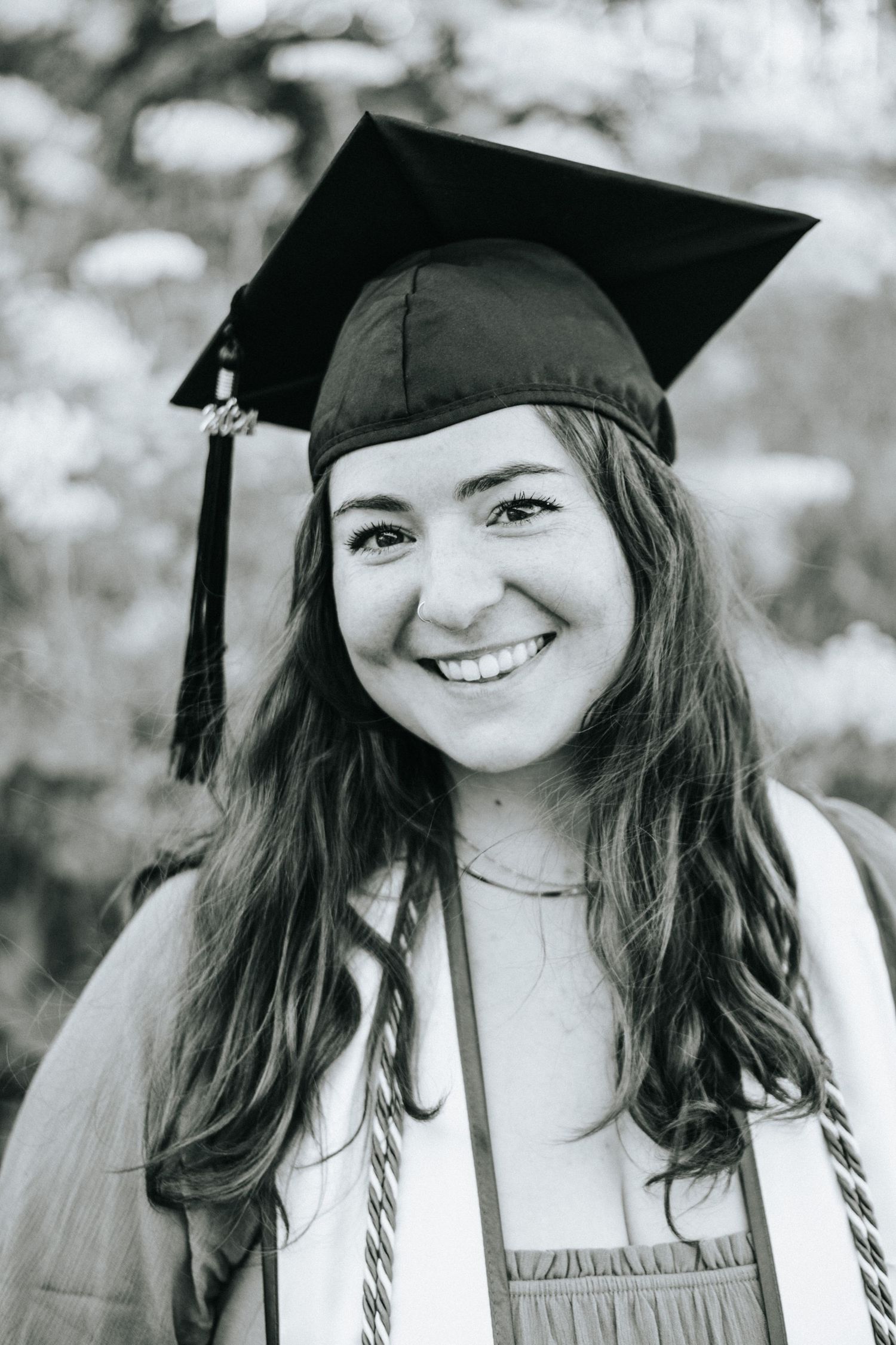 Black and white portrait of graduate in cap and gown with natural bokeh background.