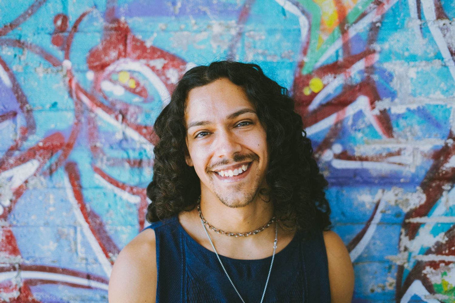 Person with long dark hair smiling in front of colorful graffiti wall.