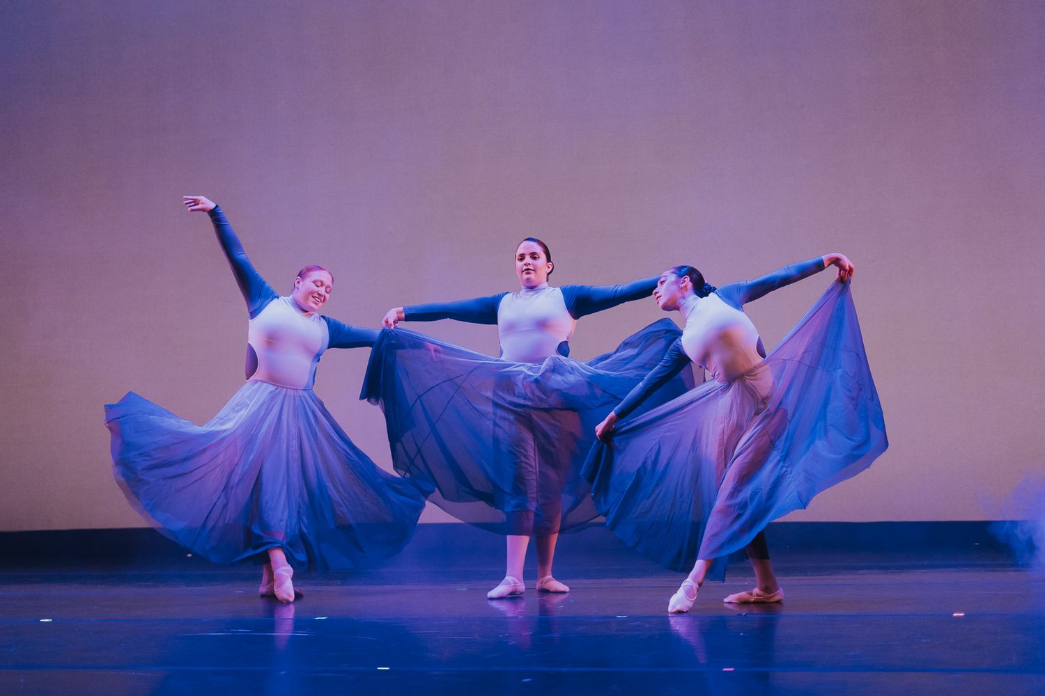 Three ballet dancers in flowing blue dresses perform synchronized movements on a dimly lit stage.