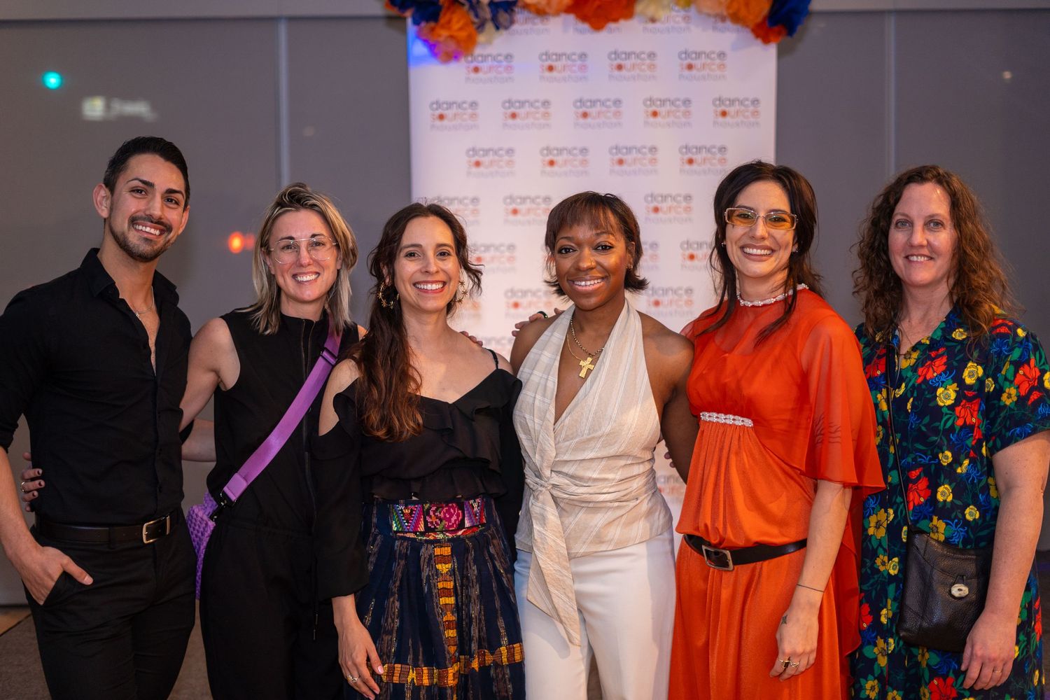 Six diverse people smiling and posing together at a formal event in front of a decorative backdrop.