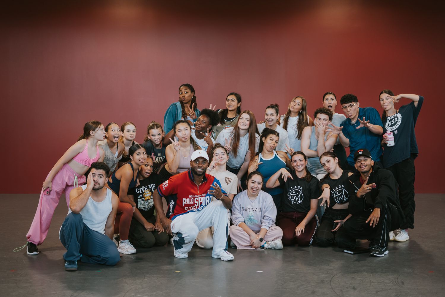 Group of dancers pose together against a red wall in casual clothing.