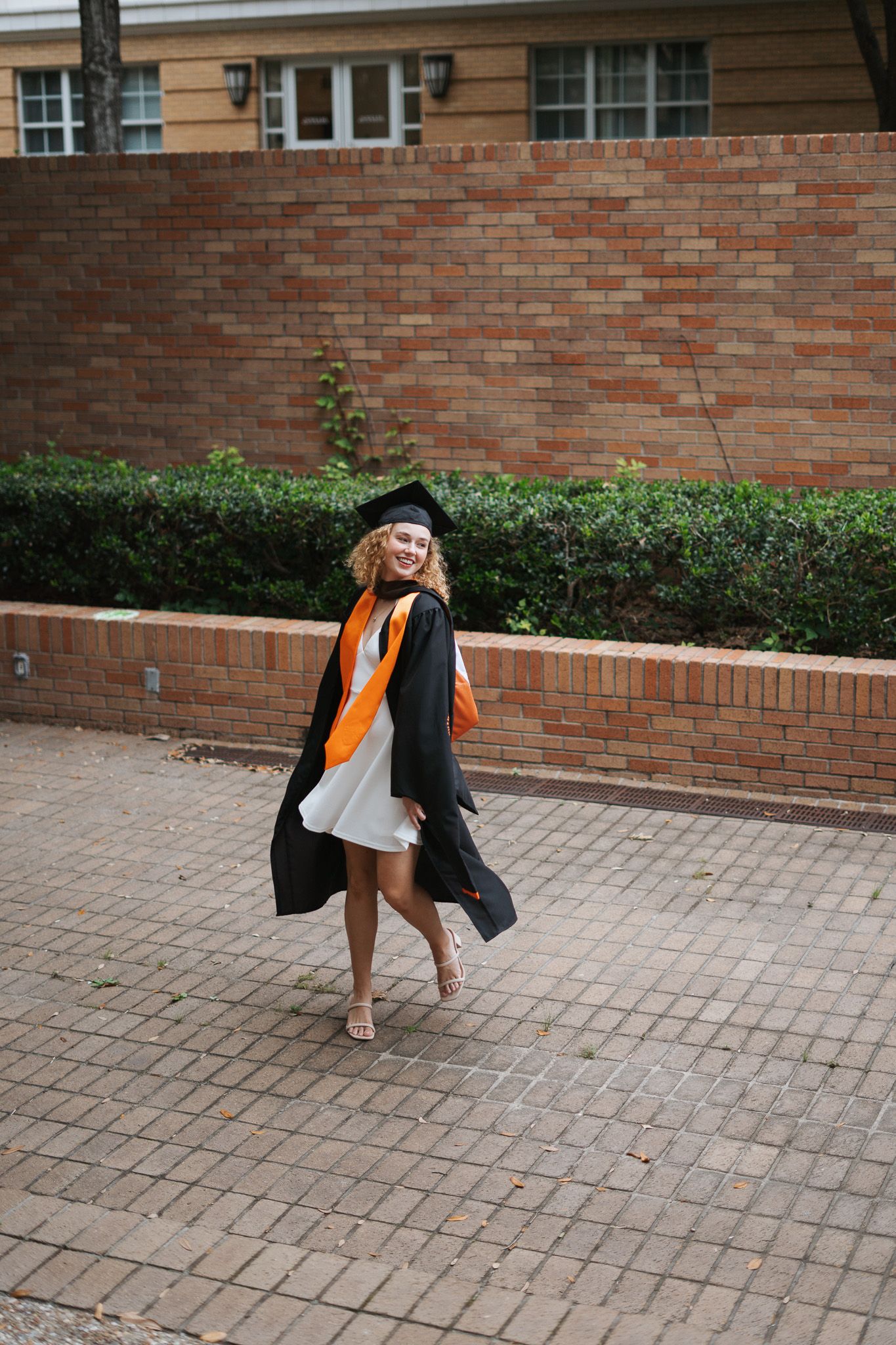 Young woman in black graduation gown and cap with orange sash walks on brick path near university building.