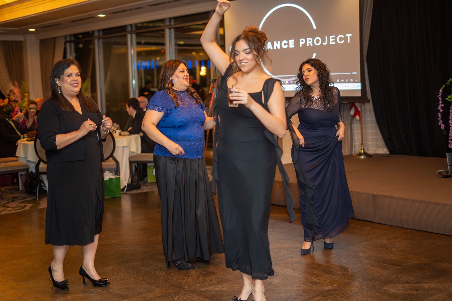 Four women in elegant black gowns dancing on stage at a Dance Project gala event.